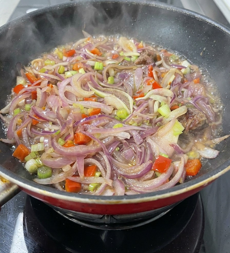 Una casseruola piena di cipolle affettate, carote, sedano e pezzi di carne che sobbollono in un liquido leggero con vapore che sale.
