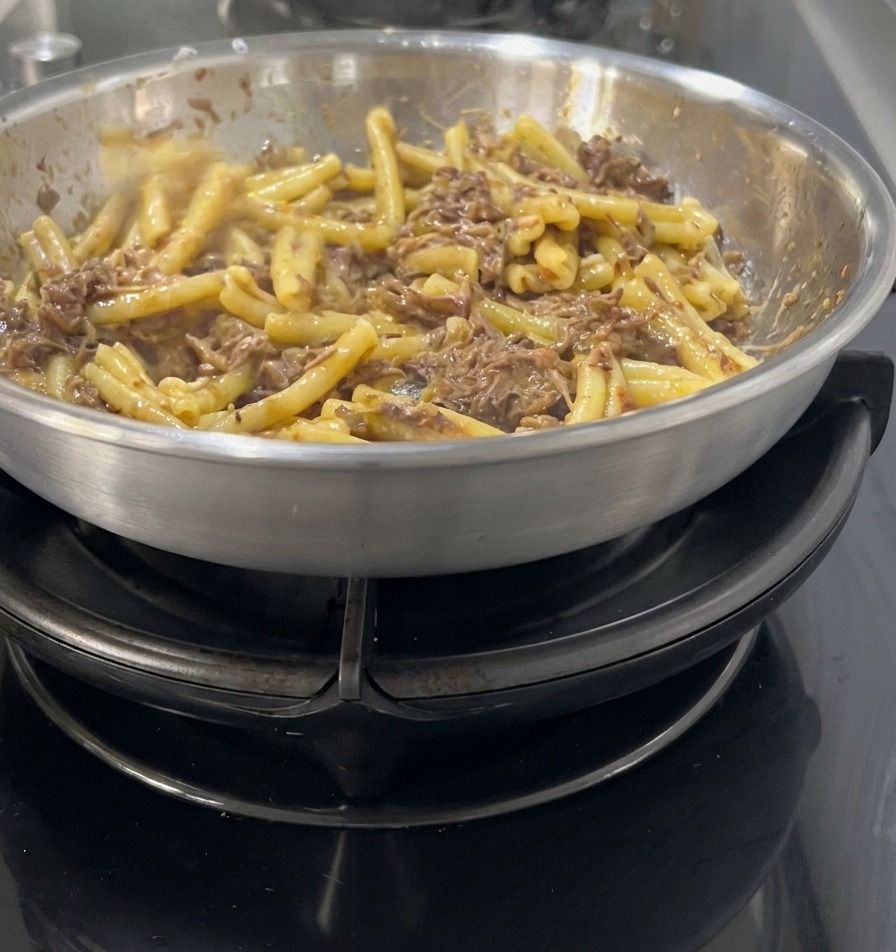 Pasta casarecce che viene saltata e amalgamata in un sugo scuro e ricco di carne sfilacciata e cipolle in una padella dacciaio.