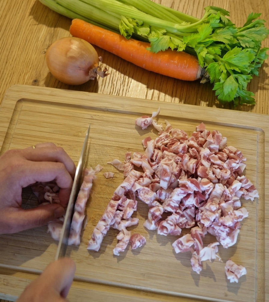 Primo piano di mani che tagliano a dadini sottili delle fette di pancetta su un tagliere di legno chiaro.