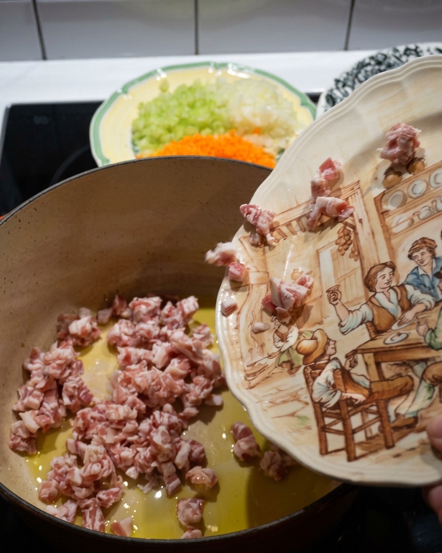 Cubetti di pancetta versati da un piatto decorato in una pentola di ghisa rossa calda con olio doliva.