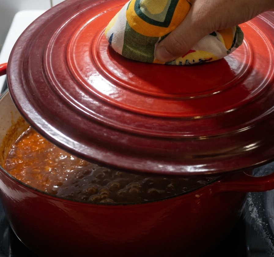 Una mano con un guanto da forno giallo e verde che posiziona un coperchio di ghisa rossa su una pentola che bolle a fuoco lento.