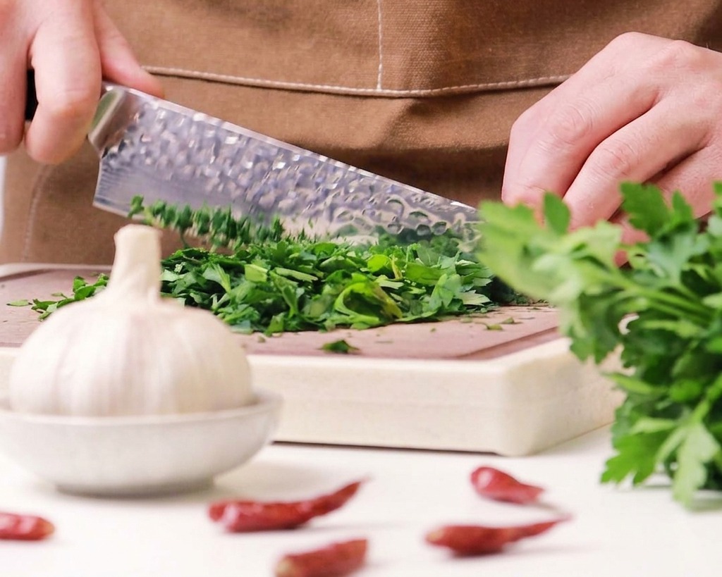 Un primo piano di uno chef che usa un coltello per tritare finemente un mucchietto di prezzemolo fresco su un tagliere di legno.