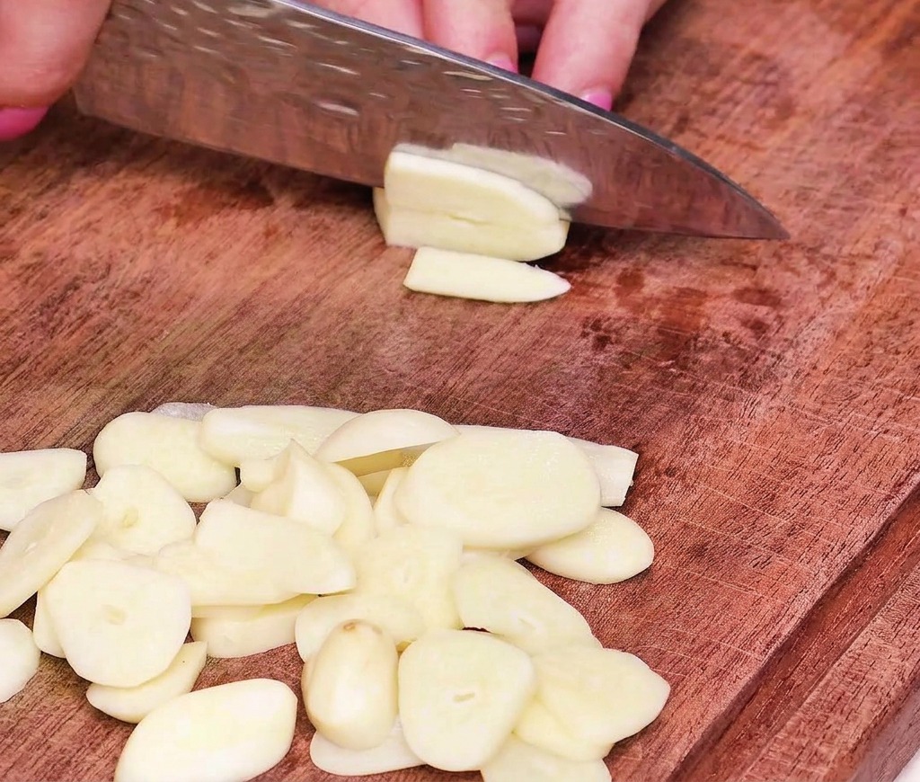 Un primo piano di una mano che usa un coltello da chef per affettare gli spicchi daglio in rondelle sottili e uniformi su un tagliere.