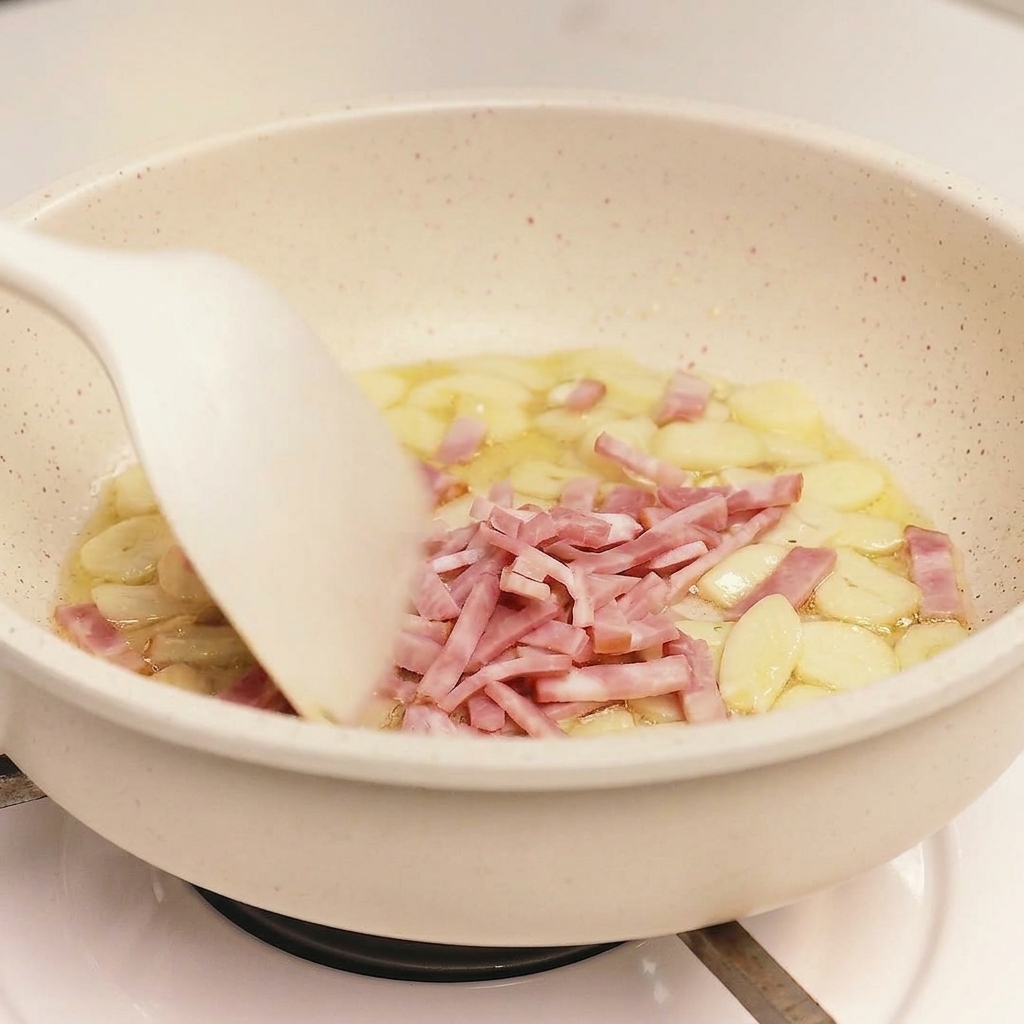Una spatola bianca mescola il bacon a pezzetti e laglio in una padella dal fondo chiaro.