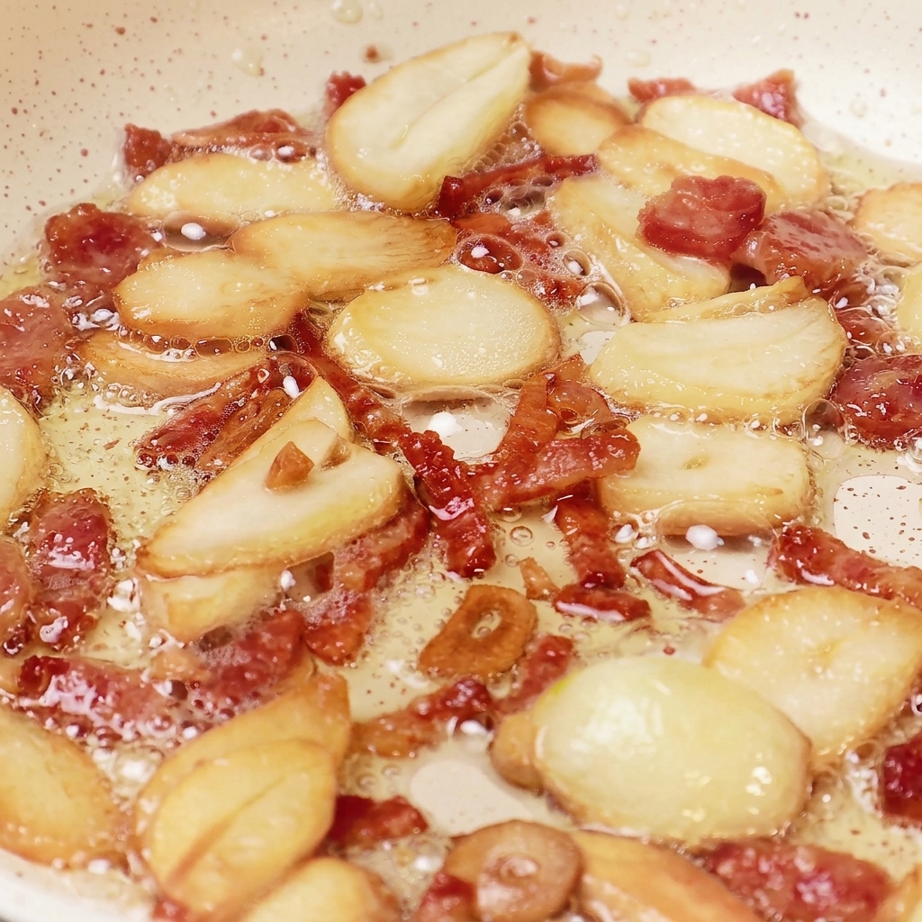 Lamelle daglio e pezzetti di bacon che sfrigolano nellolio allinterno di una padella, mostrando un colore dorato intenso e una consistenza croccante.