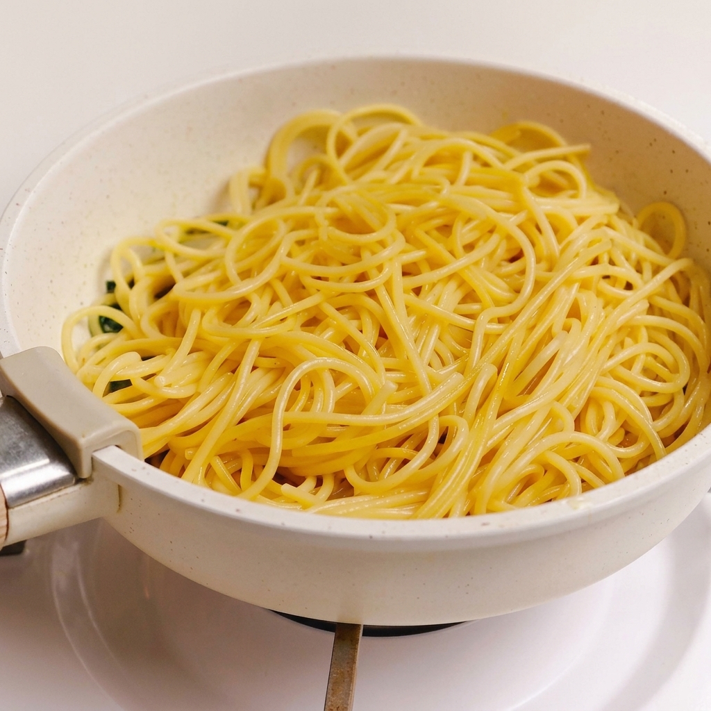 Una porzione di spaghetti cotti adagiata sopra la salsa di erbe, aglio e bacon in una padella chiara.