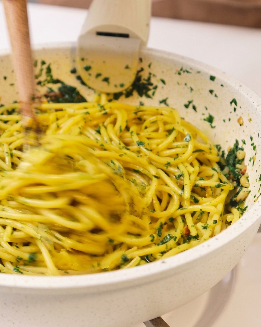 Scatto dinamico degli spaghetti che vengono saltati e mescolati con erbe e olio in padella usando un paio di bacchette di legno.
