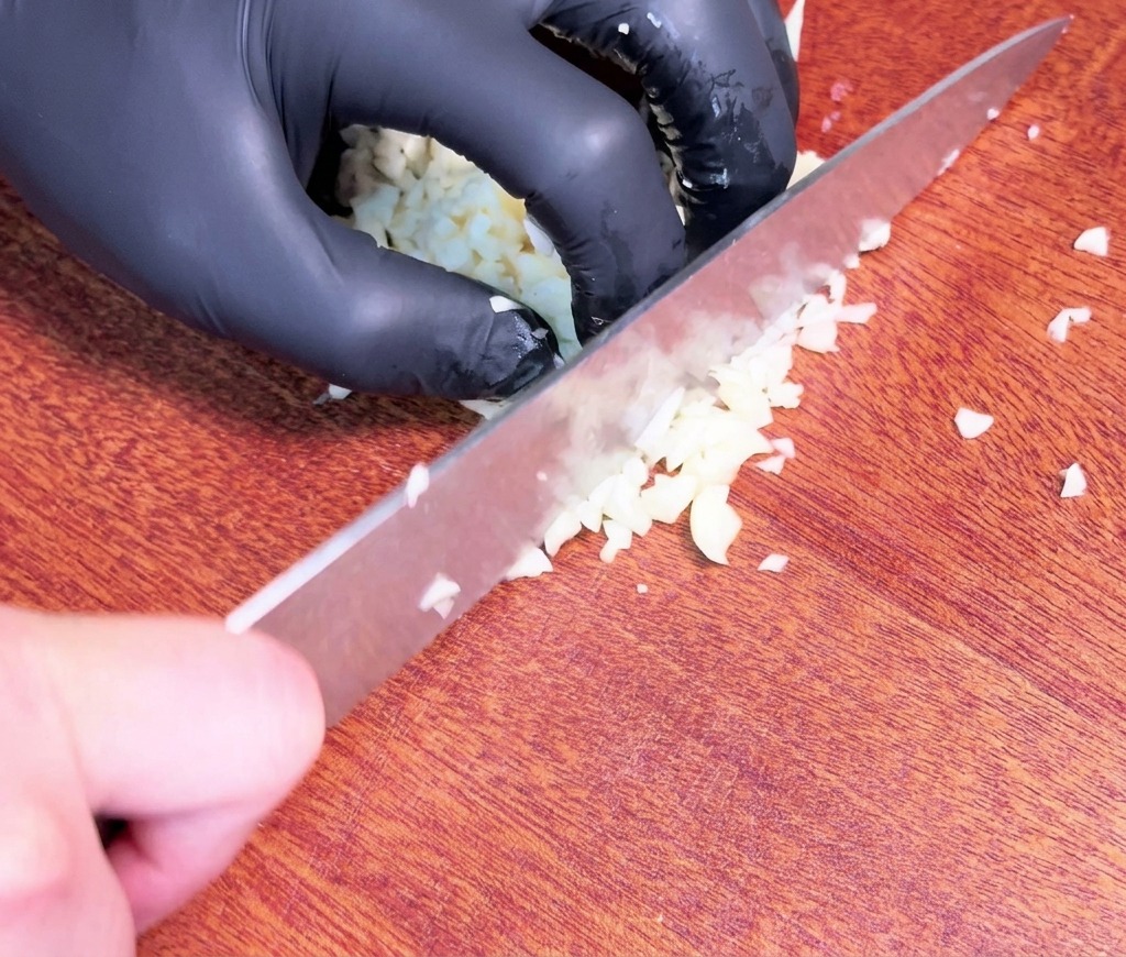 Una mano con un guanto nero usa un coltello da chef per tritare finemente gli spicchi daglio crudo su un tagliere di legno scuro.