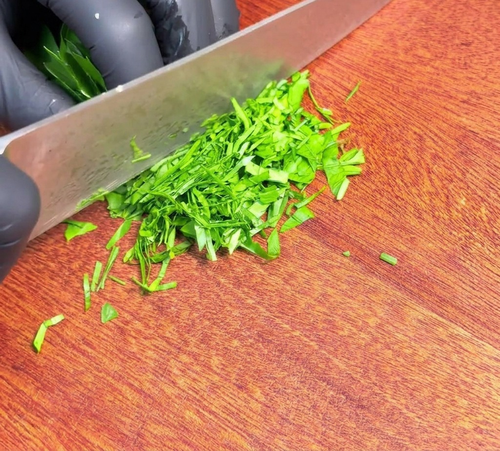 Prezzemolo fresco verde tritato finemente e distribuito su un tagliere di legno accanto alla lama di un coltello affilato.