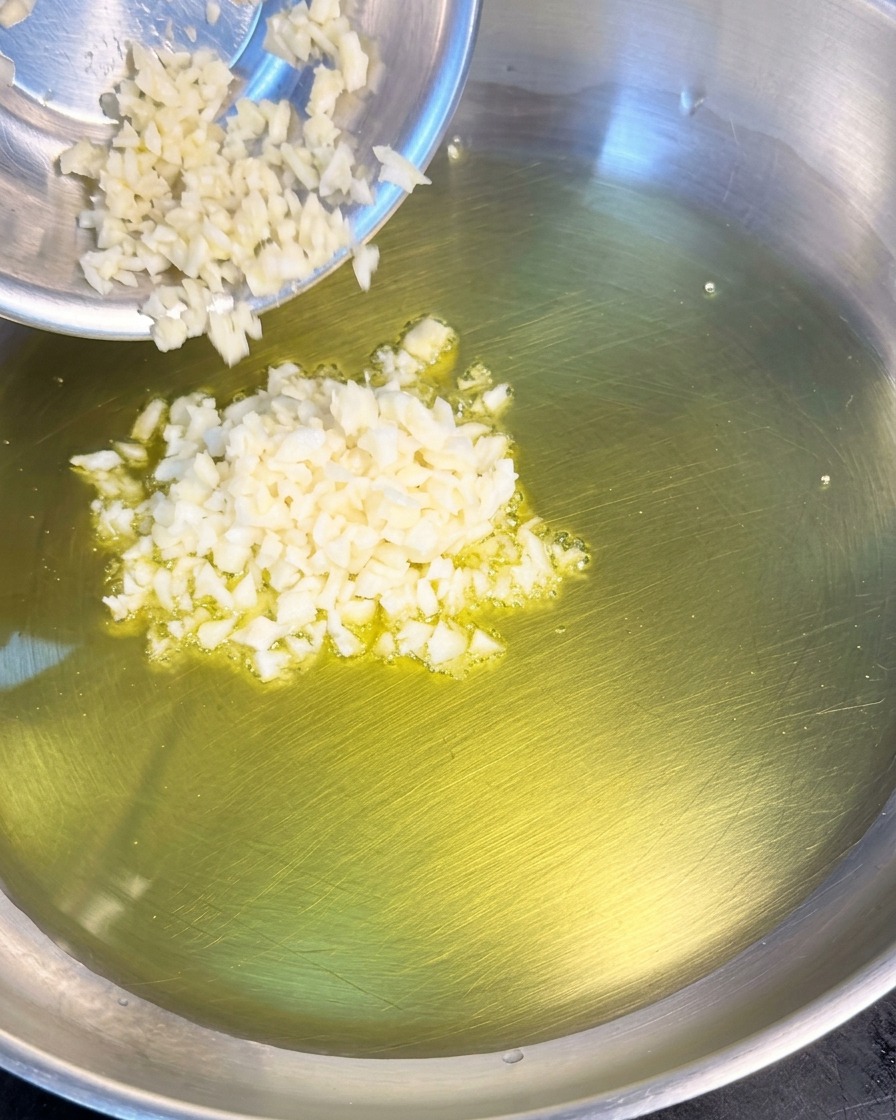 Un mucchietto di aglio tritato finemente viene aggiunto da una ciotolina di metallo in una padella con olio doliva caldo.