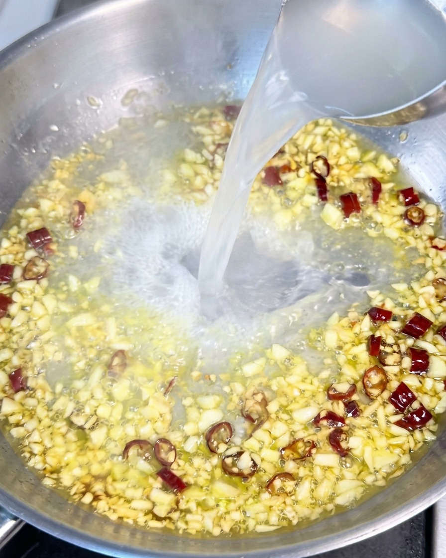 Acqua di cottura della pasta, torbida e bianca, viene versata da un mestolo in una padella con olio, aglio e peperoncino.