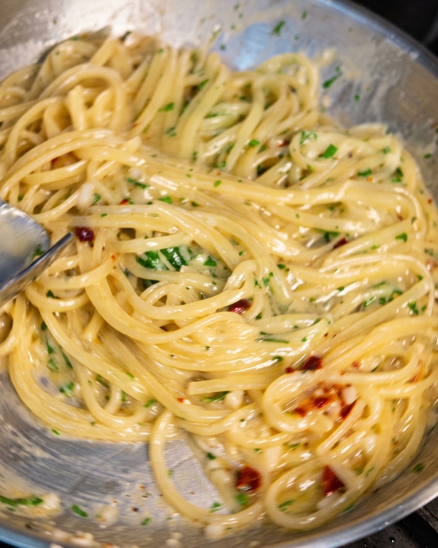 Una padella in acciaio inossidabile piena di spaghetti viene fatta saltare usando le pinze. La pasta è avvolta da una salsa lucida e cremosa con pezzetti visibili di aglio, peperoncino e prezzemolo.