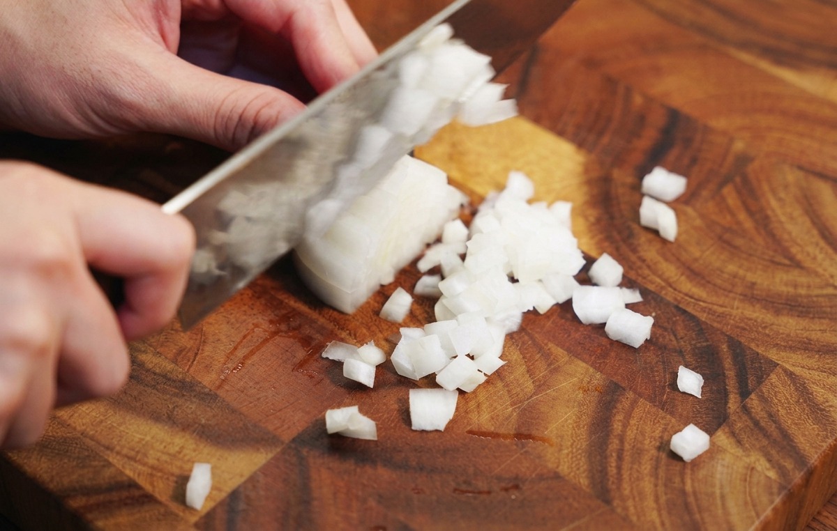 Le mani di una persona che usa un coltello da chef per tritare una cipolla bianca su un tagliere di legno.