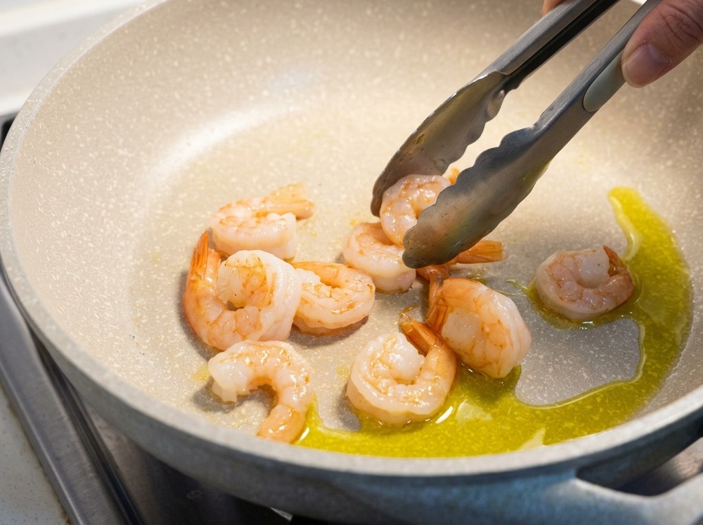 Gamberi cotti e rosati girati con pinze metalliche in una padella chiara con olio doliva caldo.