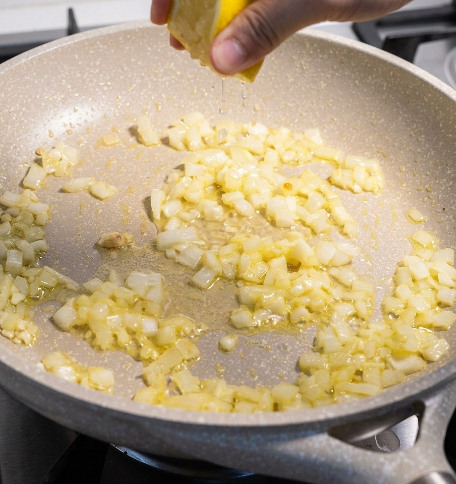 Una mano che spreme metà limone giallo su una padella contenente cipolla e aglio soffritti.