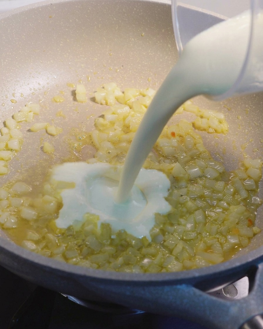 Panna da cucina versata da un misurino in una padella beige con pezzetti di cipolla e aglio soffritti.