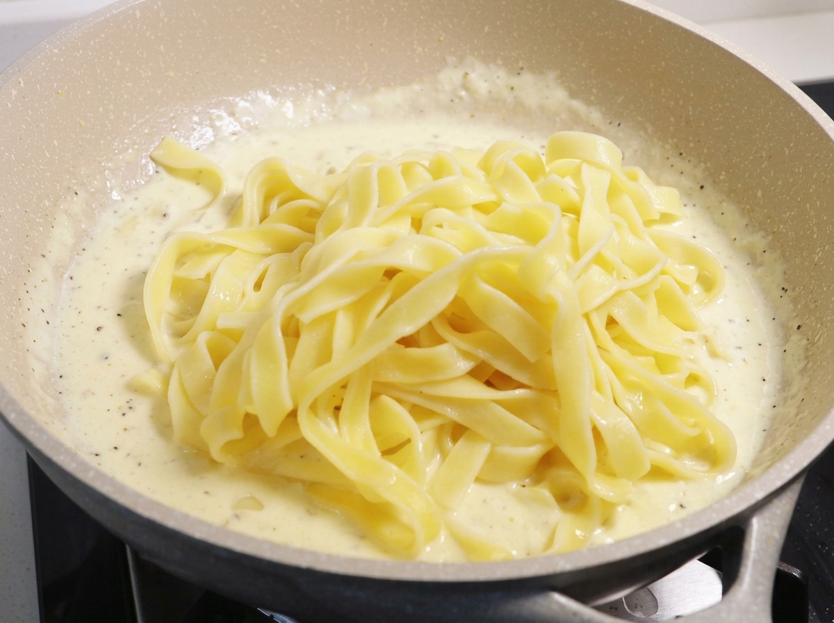 Una porzione abbondante di fettuccine gialle cotte adagiate sopra una densa salsa alla panna che sobbolle in una grande padella.