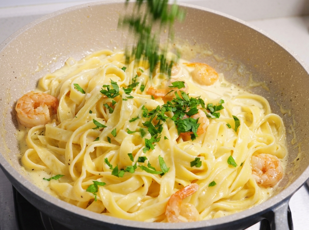 Prezzemolo verde fresco tritato cosparso su un piatto di fettuccine cremose ai gamberi in una padella chiara.