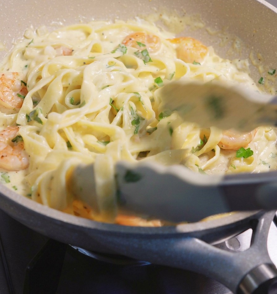 Uso delle pinze per sollevare e mescolare fettuccine e gamberi rosa in una salsa alla panna bianca allinterno di una padella.
