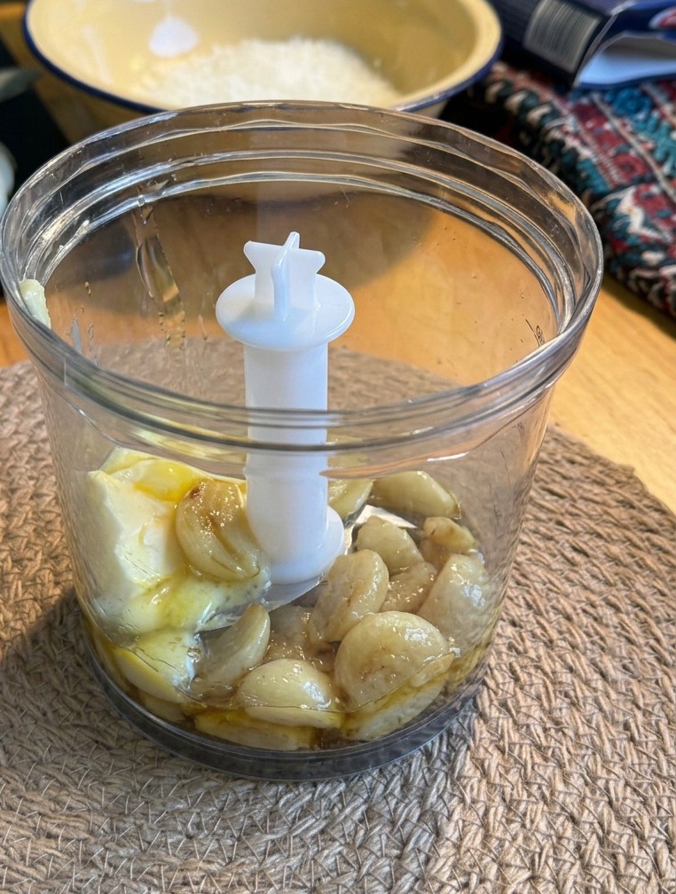 Spicchi daglio arrosto e un panetto di burro allinterno del bicchiere di un piccolo frullatore trasparente appoggiato su una tovaglietta di canapa.