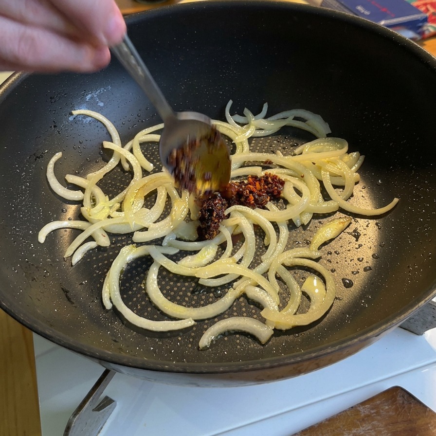 Un cucchiaio che versa del vibrante olio piccante rosso in una padella con le cipolle bianche parzialmente cotte.