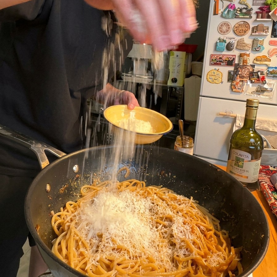 Una mano che spolvera uno strato abbondante di Parmigiano grattugiato fine sopra unampia padella di spaghetti cremosi.