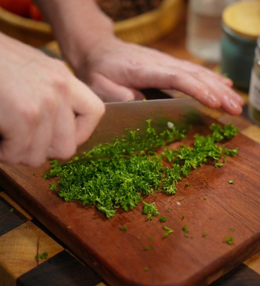Primo piano di mani che usano un grande coltello da chef per tritare finemente del prezzemolo verde brillante su un tagliere di legno scuro.