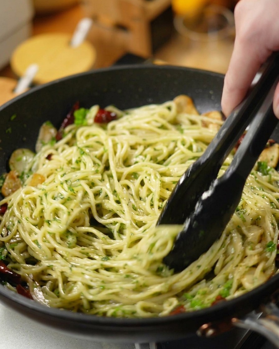 Pinze nere che saltano gli spaghetti insieme al prezzemolo tritato, aglio e peperoncino in una padella scura.