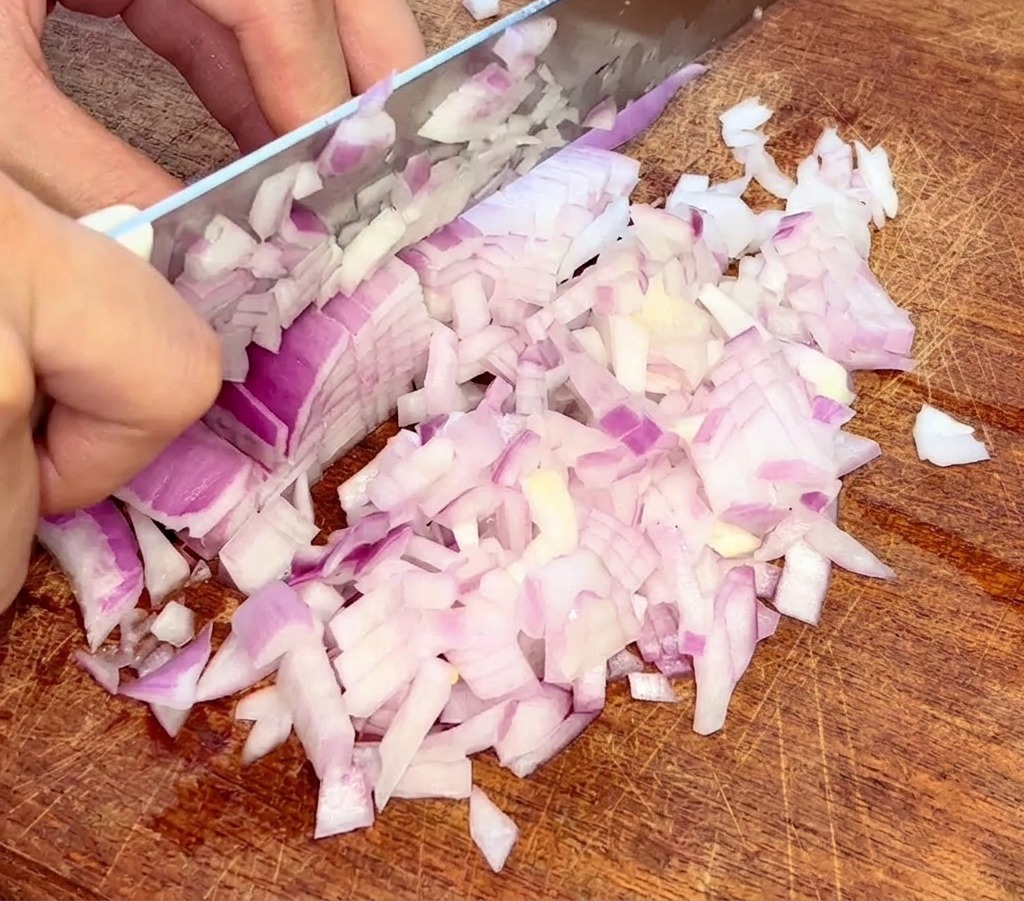 Un primo piano di mani che usano un coltello per tagliare finemente a dadini una cipolla rossa su un tagliere di legno.