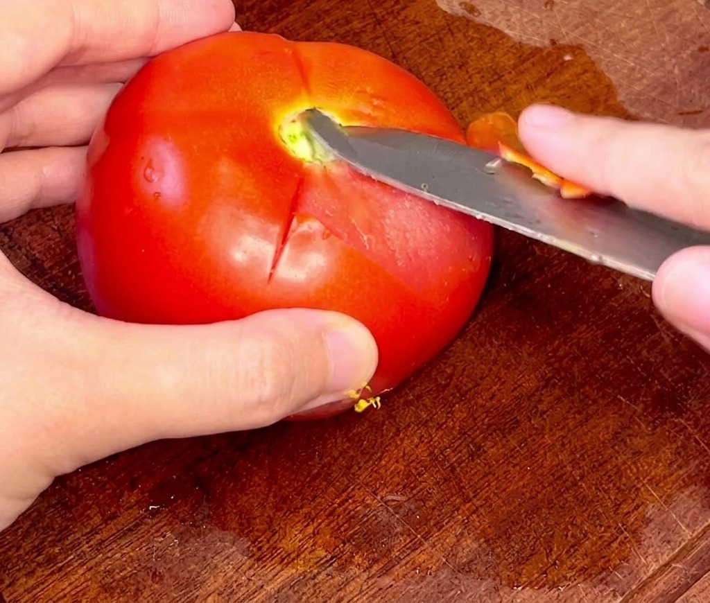Mani che usano con cura uno spelucchino per rimuovere la buccia da un pomodoro intero sbollentato.