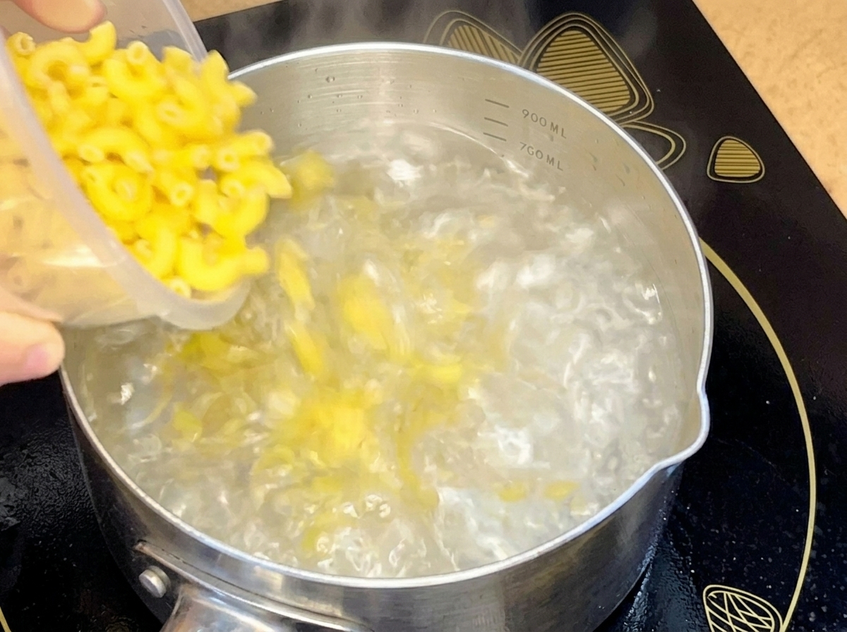 Maccheroni crudi versati da un contenitore in una pentola di acciaio inossidabile con acqua bollente.