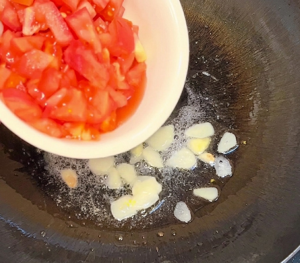 Pomodori a dadini versati da una ciotola bianca in una padella con olio caldo e fettine daglio dorate.