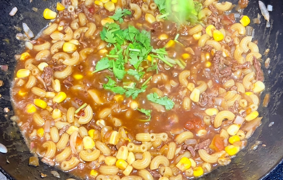 Una mano spolvera del coriandolo verde tritato su una padella fumante di maccheroni con manzo e pomodoro.
