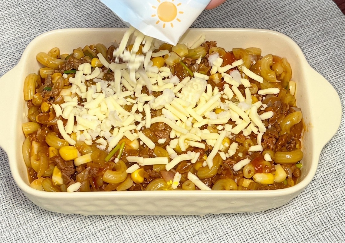 Formaggio bianco grattugiato versato da una confezione blu e bianca su una pirofila piena di maccheroni al sugo.