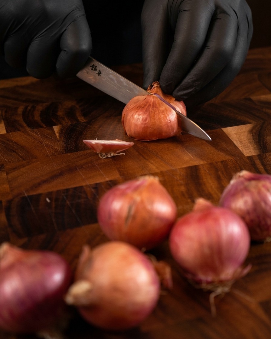 Mani con guanti neri che usano un coltello affilato per affettare finemente lo scalogno su un tagliere di legno scuro.