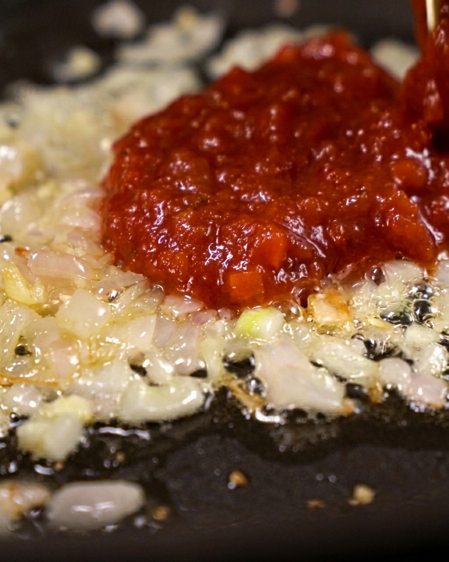 Un generoso cucchiaio di concentrato di pomodoro rosso vivo aggiunto sopra lo scalogno appassito in padella.
