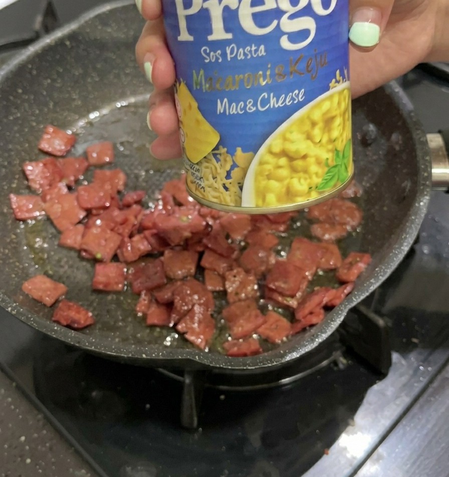 Una mano che tiene una lattina di salsa per Mac e Cheese sopra la padella con il salame rosolato.