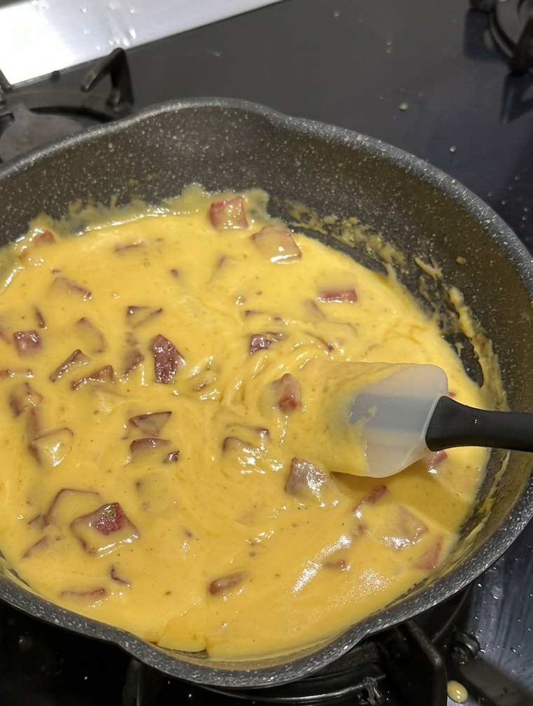 Una padella antiaderente nera piena di maccheroni, salame a cubetti e una densa salsa gialla al formaggio, mescolati con una spatola di silicone bianca.