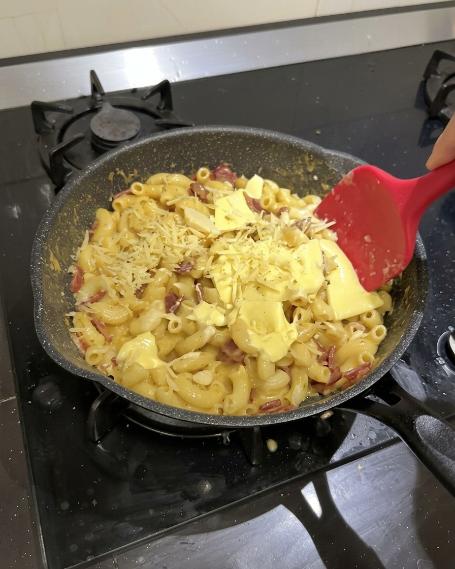 Maccheroni, salame e formaggio a fette in una padella nera mentre vengono mescolati con una spatola di silicone rossa.