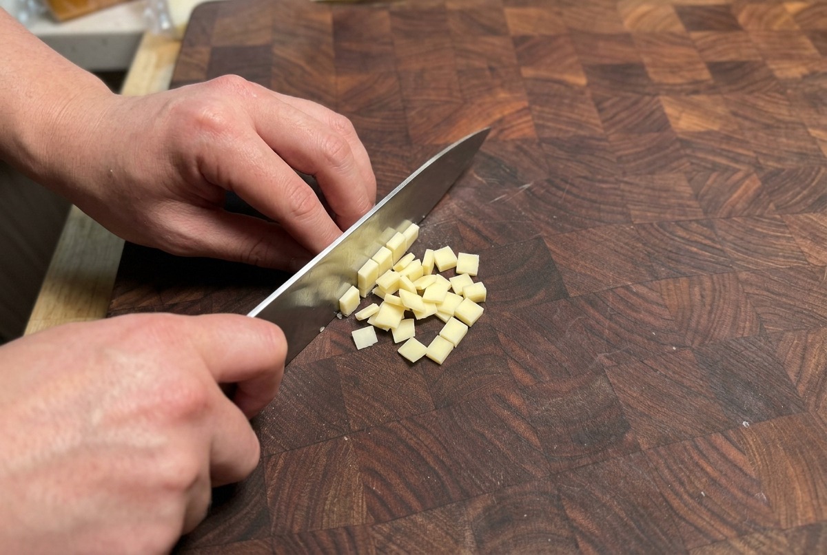 Mani che usano un coltello da chef per tagliare a cubetti un blocco di formaggio giallo chiaro su un tagliere di legno.
