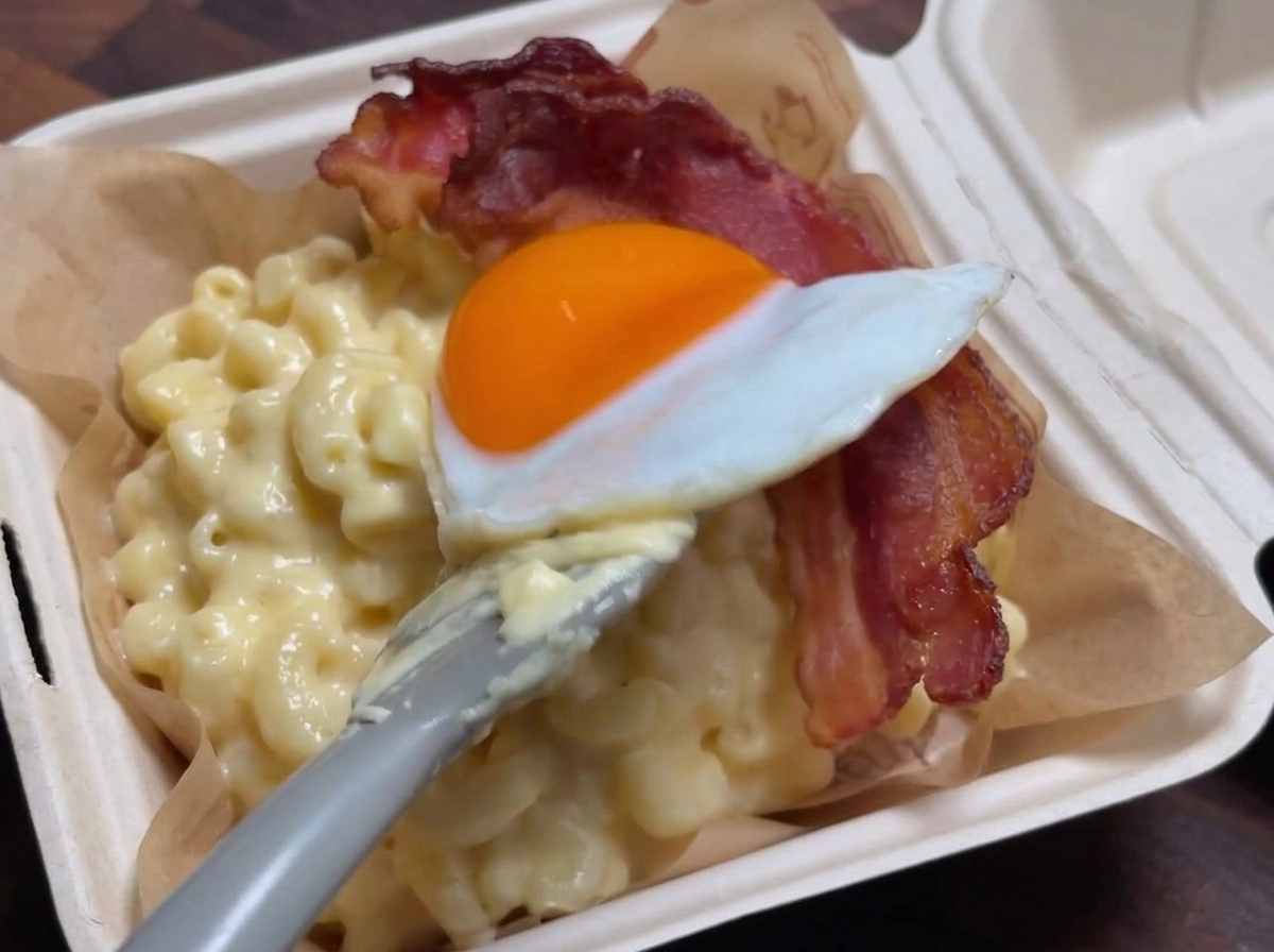 Una spatola che adagia delicatamente un uovo allocchio di bue sopra una striscia di bacon croccante adagiata su una porzione di maccheroni al formaggio.