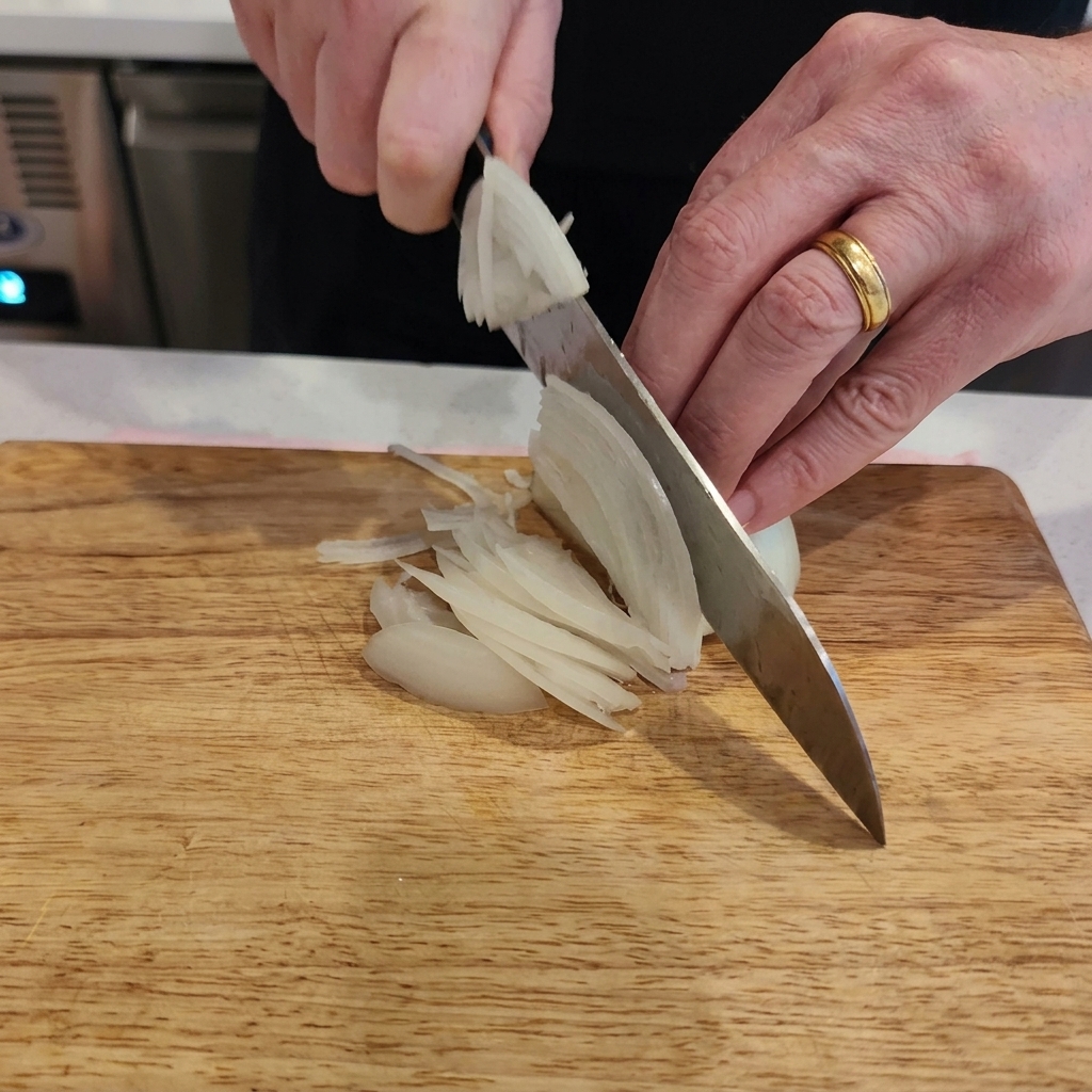 Un primo piano delle mani di una persona che usa un coltello da chef per tagliare una cipolla bianca a strisce sottili su un tagliere di legno chiaro.