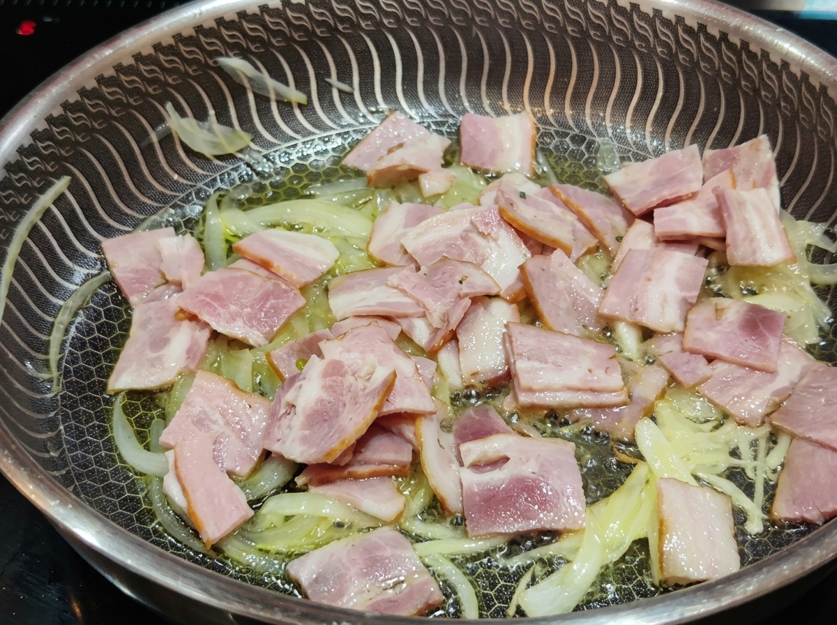 Fette di pancetta rosa che cuociono in padella insieme a strisce di cipolla ammorbidite e traslucide nellolio.
