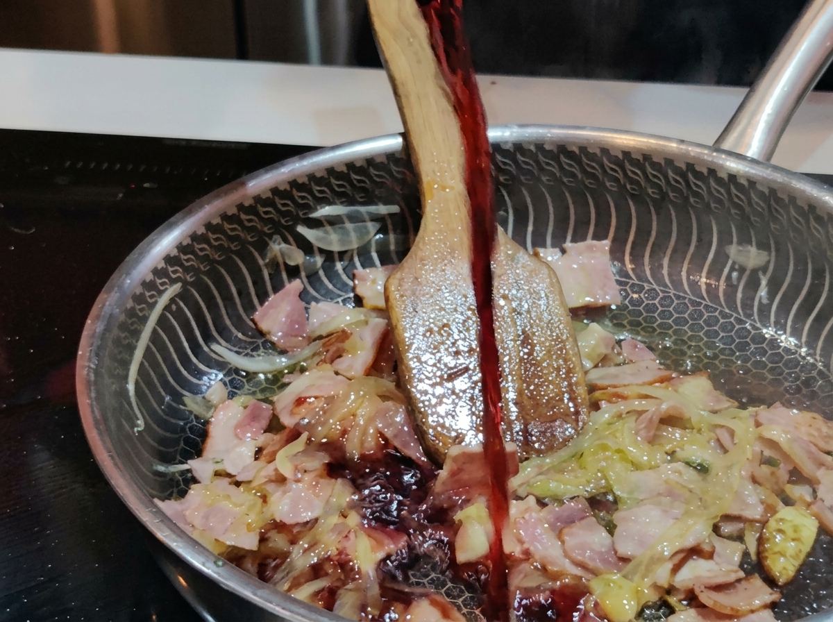 Un filo di vino rosso scuro versato da un bicchiere in una padella in acciaio inossidabile piena di pancetta e cipolle sfrigolanti.