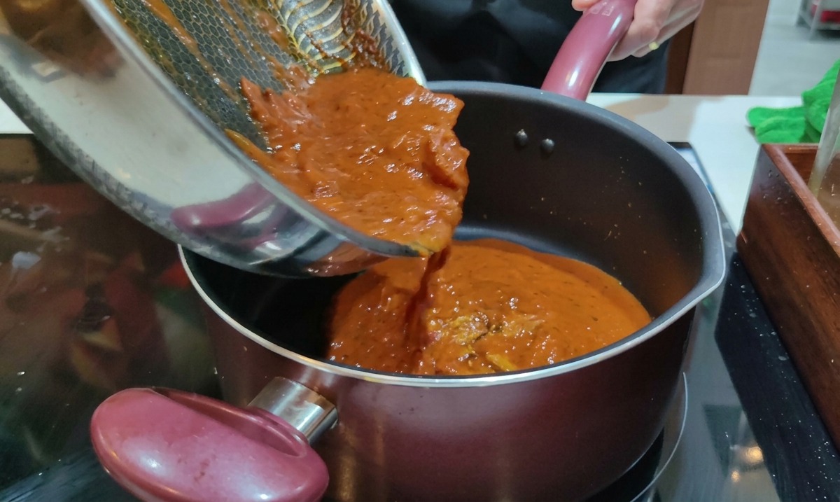 Sugo rosso allAmatriciana appena cotto versato da una padella in una pentola di rigatoni bolliti.