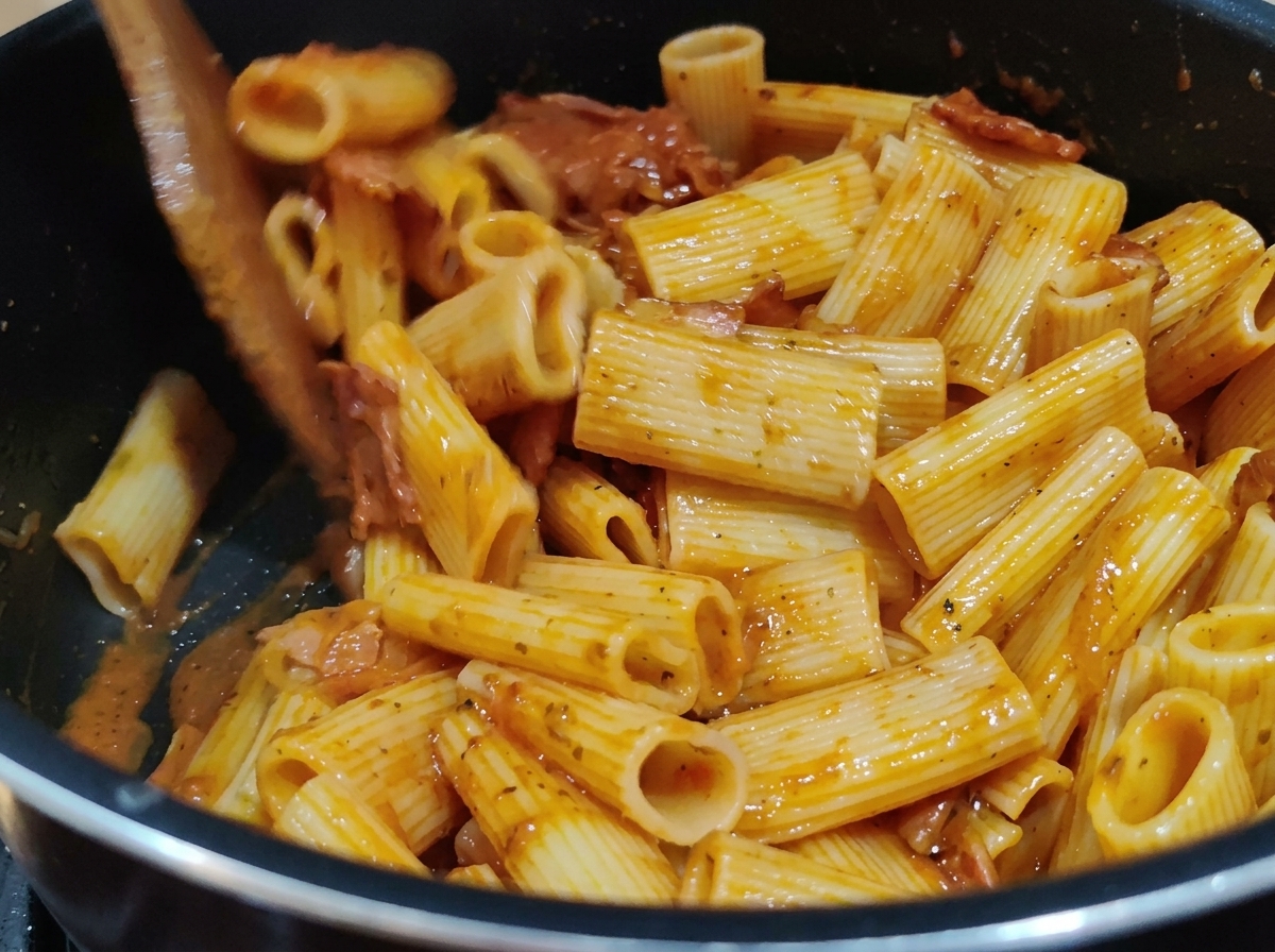 Rigatoni mescolati con un ricco sugo di pomodoro rosso in una pentola usando una spatola di legno.