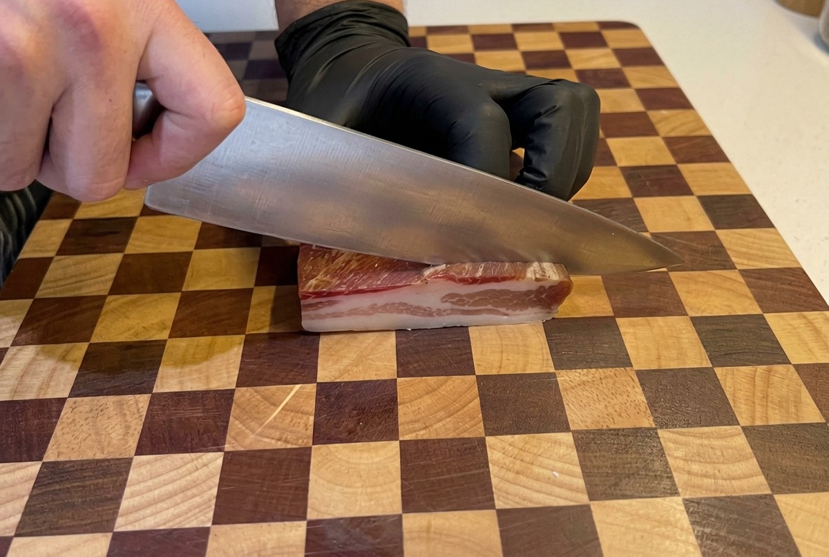 Mani con guanti neri usano un coltello da chef per tagliare una fetta spessa di guanciale a cubetti su un tagliere di legno.