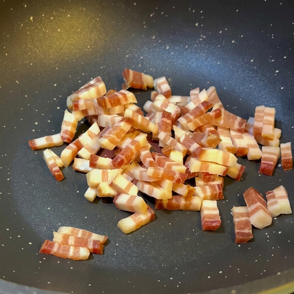 Cubetti di pancetta italiana che rosolano in una padella antiaderente scura, mostrando il grasso sciolto e una leggera doratura sui bordi.