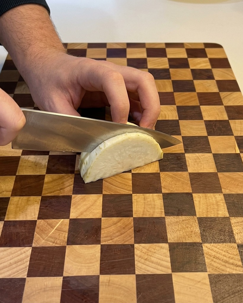 Una persona usa un coltello da chef per tritare finemente uno spicchio di cipolla bianca su un tagliere di legno a scacchi.