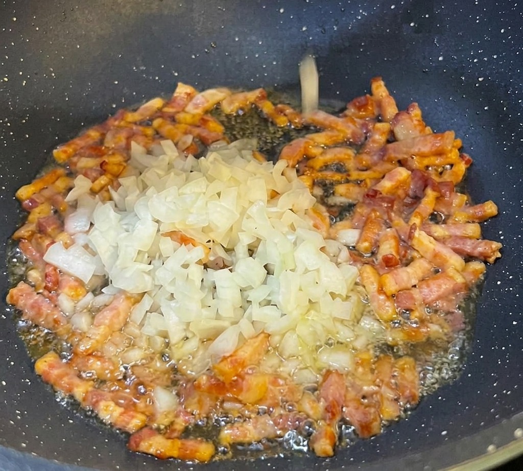 Cipolla bianca tritata finemente che viene aggiunta in una padella nera contenente striscioline di pancetta ben dorate.